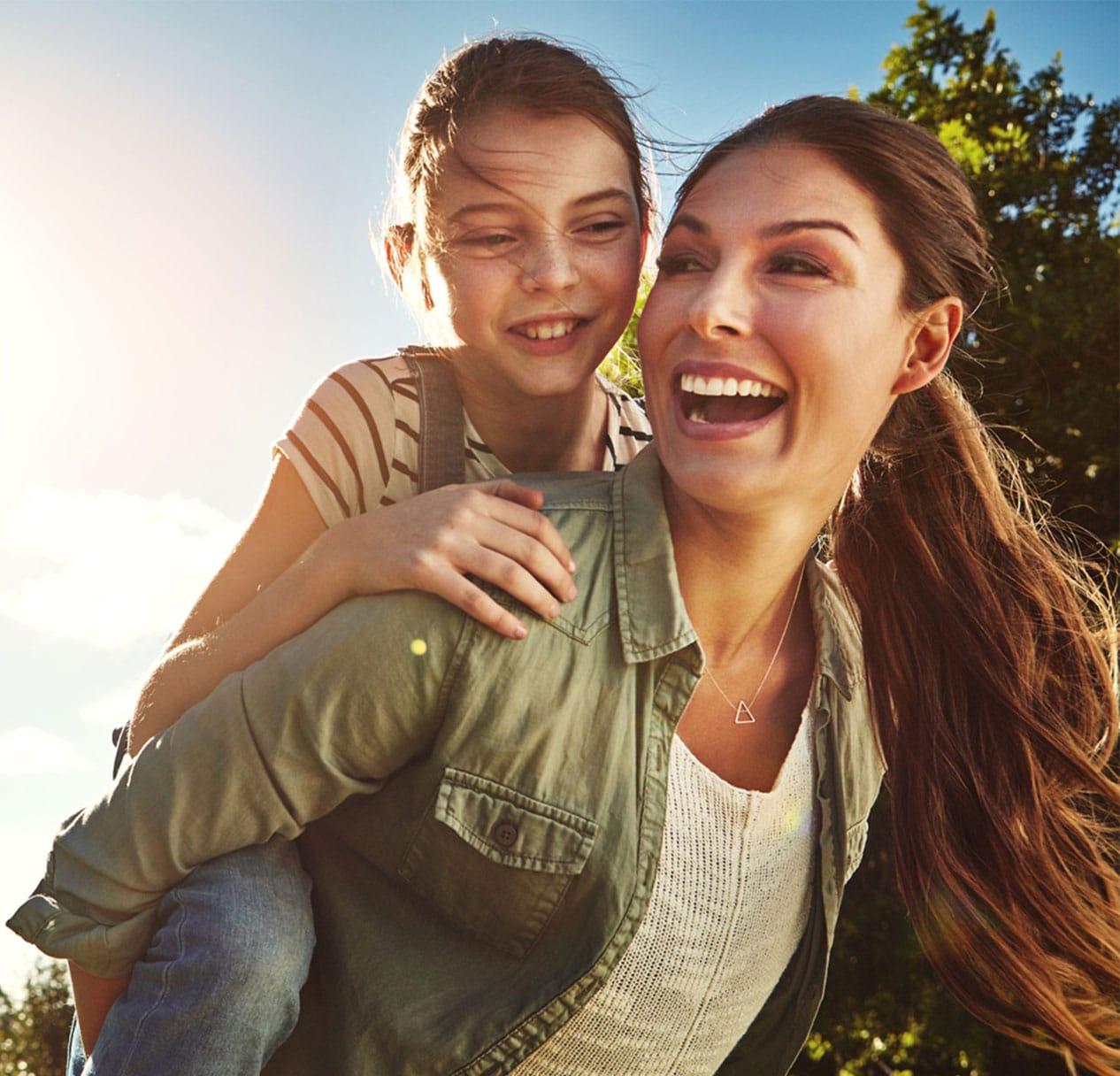 Smiling woman giving her young daughter a piggyback ride in the sunshine – Craig Family Dental