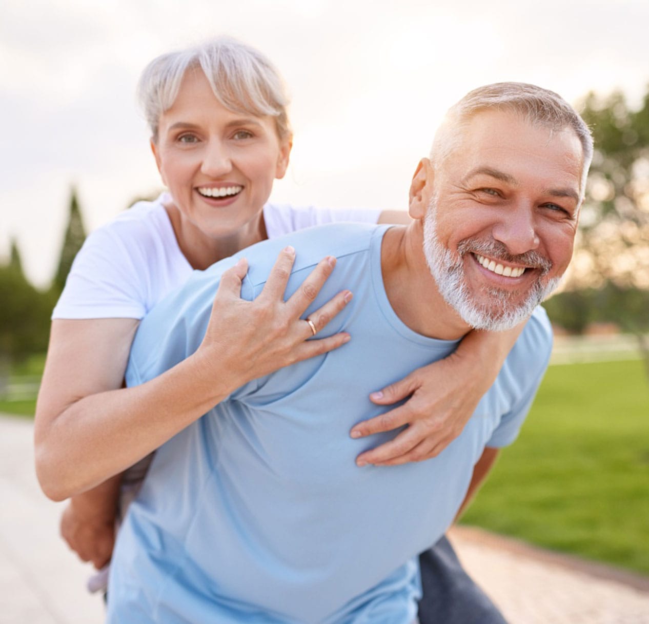 Smiling older couple enjoying a piggyback ride in a sunny park – Craig Family Dental